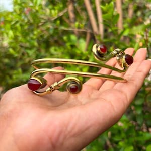 Può includere: Un bracciale a manetta in metallo color oro con un design a doppio filo e terminali a spirale. Ognuna delle quattro estremità è impreziosita da una pietra preziosa liscia rosso scuro, a forma di goccia o rotonda, per un aspetto simmetrico.