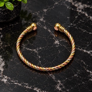 May include: A tri-color metal cuff bracelet featuring a twisted rope design made of intertwined gold, silver, and copper-toned strands. The open-ended bracelet terminates in two polished spherical end caps with a smooth, reflective finish.