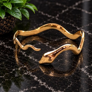 Op de afbeelding: Een goudkleurige metalen armband in de vorm van een kronkelende slang met een gladde, gepolijste afwerking. Het open ontwerp heeft een gedetailleerde kop aan het ene uiteinde en een toelopende staart aan het andere, met een golvend lichaam.