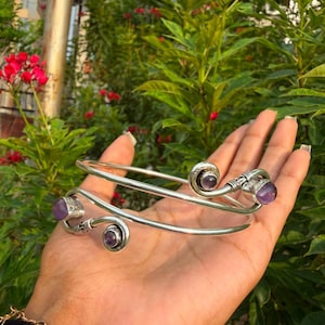 Puede incluir: Un brazalete de metal en tono plateado con un diseño en espiral y detalles de piedras preciosas moradas pulidas en cada extremo. El brazalete tiene una banda lisa y redondeada que se enrolla, mostrando dos piedras moradas en engastes de metal.