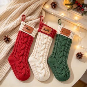 May include: Three cable-knit Christmas stockings in red, cream, and dark green. Each stocking features a ribbed cuff with a rectangular brown leather patch attached by four metal rivets. The names Thea, Kylie, and Olive are printed on the patches.