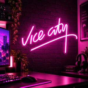 Op de afbeelding: Een felroze neonbord gemonteerd op een donkere bakstenen muur met de tekst Vice city in een cursief lettertype. Het bord straalt een levendige gloed uit die de omgeving verlicht, waaronder een bureau met een computermonitor en een koptelefoon.