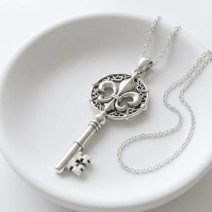 May include: A silver-toned metal key pendant necklace featuring a fleur-de-lis emblem centered within a circular filigree frame. The key has a textured shaft and a standard bit, suspended from a delicate silver-toned cable chain link necklace.