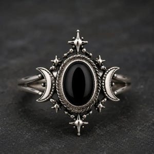 May include: A silver-toned ring featuring a central oval black stone surrounded by a twisted rope border. The setting is accented with two crescent moon shapes on the sides and four small star-shaped points at the top and bottom.