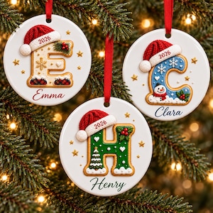 May include: Three round white ceramic ornaments hanging by red ribbons. Each features a large letter E, C, or H decorated like a cookie with a red Santa hat labeled 2026. The names Emma, Clara, and Henry are printed below the letters.