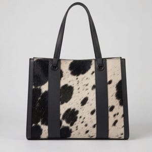 Könnte beinhalten: Eine strukturierte Tragetasche mit einem schwarz-weißen Kuhfellmuster auf der Vorder- und Rückseite. Die Tasche verfügt über zwei schwarze Ledergriffe, schwarze Lederseitenteile und vertikale schwarze Lederriemen mit runden Beschlägen.