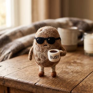 May include: A brown needle-felted potato figure with a textured surface, wearing black sunglasses. The figure has two small arms and legs, and is holding a small white mug with the word BUSY written in black letters on the front of the mug.