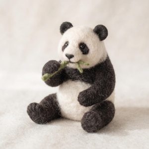 Op de afbeelding: Een met de naald gevilt panda-figuurtje van wol, met een wit gezicht en buik en zwarte oren, ledematen en oogvlekken. De panda zit in een zittende houding en houdt een kleine, lichtgroene gevilde bamboestengel bij zijn mond vast.