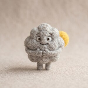 May include: A needle-felted grey cloud figurine with a textured, woolly surface, featuring two large white eyes with black pupils, a small smile, two small arms held against its chest, and a bright yellow sun shape attached to its side.