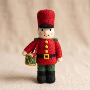Può includere: Una statuetta di soldatino in feltro ad ago che indossa un cappello rosso alto, una giacca rossa con tre bottoni gialli, una cintura nera con fibbia gialla, pantaloni verdi e stivali neri. La figura tiene in mano un piccolo tamburo verde.