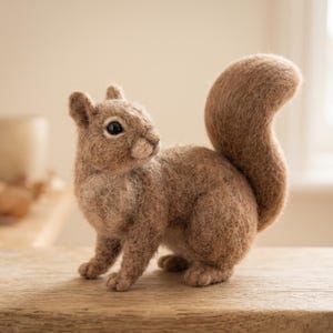 Könnte beinhalten: Eine nadelgefilzte Eichhörnchenfigur aus hellbraunen Wollfasern. Das Eichhörnchen hat ein strukturiertes, flauschiges Fell, einen großen geschwungenen Schwanz, kleine spitze Ohren und dunkle, runde Augen und steht in einer aufrechten Haltung.