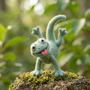Puede incluir: Una figura de lagarto de lana fieltrada de color verde claro con una superficie texturizada y difusa. El lagarto tiene ojos grandes y redondos, una sonrisa amplia y una pequeña lengua rosa que sobresale. Está posado con la cola curvada hacia arriba.