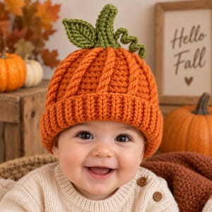 May include: A bright orange crochet beanie designed to resemble a pumpkin, featuring a ribbed brim, a textured cable knit pattern, and a green stem with a leaf and a curly vine attached to the top of the hat, worn by a smiling infant.