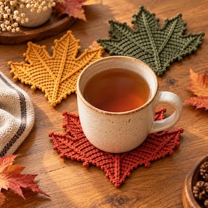 May include: Three crocheted leaf-shaped coasters in mustard yellow, olive green, and burnt orange. A speckled cream-coloured ceramic mug filled with tea sits on the orange coaster. The coasters feature intricate, raised vein-like textures.