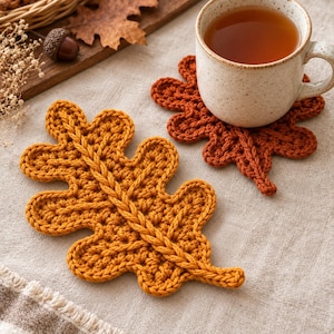 May include: Two crochet coasters shaped like oak leaves, one in a vibrant mustard yellow and the other in a deep burnt orange. The coasters feature a textured stitch pattern with a prominent central vein detail, designed to hold a ceramic mug.
