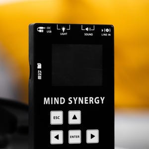 Könnte beinhalten: Ein schwarzes, rechteckiges elektronisches Gerät mit der Aufschrift MIND SYNERGY, einem zentralen Bildschirm und einem Tastenfeld mit Richtungspfeilen, einer ESC-Taste und einer ENTER-Taste. Oben befinden sich Symbole für DC USB, Licht, Ton und Line-In.