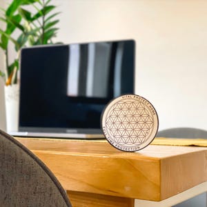 Puede incluir: Un disco circular de madera con un patrón geométrico de la flor de la vida grabado con láser. El texto VITAL PROTECT HOME y FULL TIME PROTECTION está impreso a lo largo del borde exterior del disco en una fuente negra sin remates.