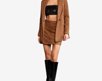 Op de afbeelding: Een lichtbruine blazer en bijpassende minirok, gecombineerd met een zwarte strapless top en zwarte kniehoge laarzen. De blazer heeft een reverskraag en twee knopen, terwijl de rok een gestructureerde tailleband en een rechte zoom heeft.