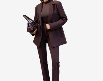 Op de afbeelding: Een donkerbruin pak bestaande uit een gestructureerde blazer, een bijpassende top met hoge hals en een pantalon. De outfit is gecombineerd met donkerbruine pumps met een puntige neus en een handtas. Grote vierkante zonnebril met donkere glazen.