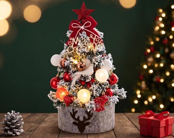 Op de afbeelding: Een kleine kunstkerstboom met besneeuwde takken, versierd met rode en witte kerstballen, een witte vogel en een rode strik. Een gouden bordje toont de tekst Merry Christmas. De boom staat in een grijze cilindervormige voet met een hert.