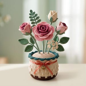 Puede incluir: Un arreglo floral de ganchillo en una maceta beige texturizada con base marrón oscuro y borde festoneado en color verde azulado. La maceta está decorada con una cinta fina color melocotón y un pequeño dije de corazón dorado.