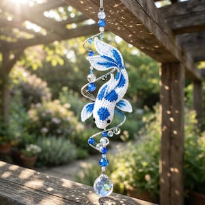 May include: A hanging mobile featuring a crocheted white and blue koi fish suspended within a silver-toned metal spiral. The ornament is decorated with clear and blue glass beads, ending with a faceted crystal sphere at the bottom.