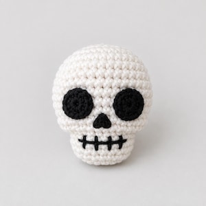 May include: A small, round, crocheted skull figure made with off-white yarn. It features two large, circular black yarn eye sockets, a small black triangular nose, and a horizontal black stitched line representing the mouth with vertical teeth.