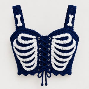 Op de afbeelding: Een donkerblauwe gehaakte crop top met een wit ribbenkastpatroon aan de voorzijde en een centrale vetersluiting met zilverkleurige metalen oogjes. De schouderbandjes zijn aan beide kanten versierd met een klein wit botmotief.