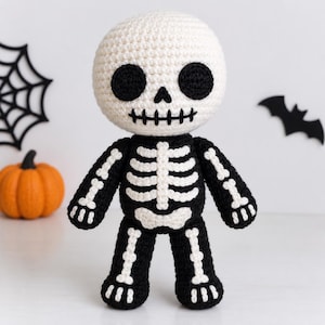 May include: A crocheted skeleton doll featuring a large, round off-white head with black embroidered eyes, a triangular nose, and a stitched mouth. The body is black with white crocheted rib cage, pelvic bone, arm, and leg bone details.
