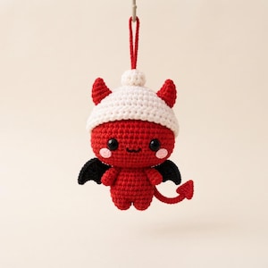 Può includere: Un ornamento a forma di diavolo all'uncinetto rosso con un berretto bianco, ali nere e una coda a punta. La figura ha due piccole corna, occhi neri e guance rosa. Un cappio di filo rosso è fissato sulla parte superiore del berretto per appenderlo.