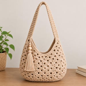 May include: A beige crochet hobo bag featuring a floral granny square pattern, a single shoulder strap, and a decorative tassel accent with a wooden bead. The bag is constructed from thick yarn with an openwork design and a solid base.