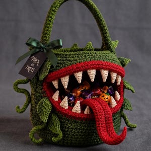 May include: A green crocheted basket shaped like a monster with a wide mouth, red lips, and cream-colored teeth. A long red tongue extends from the mouth, which is filled with candy. A dark green ribbon and a tag reading Trick or Treat are attached.