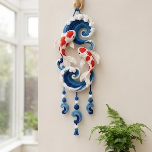 Può includere: Una decorazione da parete all'uncinetto con due pesci koi rossi, bianchi e arancioni che nuotano tra onde all'uncinetto blu e bianche. L'oggetto presenta tre fili pendenti con piccole sfere all'uncinetto blu e bianche e dettagli a forma di onda.