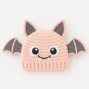May include: A light pink crochet hat featuring a bat design with dark gray ears and wings. The hat has two large white and black circular eyes, a black stitched mouth, and two small white fangs centered below the eyes on the front panel.