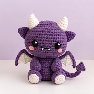 Könnte beinhalten: Ein violettes Häkel-Plüschtier in Form eines kleinen Drachen mit zwei weißen Hörnern, weißen Flügelakzenten und einem langen Schwanz mit herzförmiger Spitze. Die Figur hat große schwarze Augen, zwei kleine weiße Fangzähne und rosa Wangen.