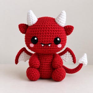 May include: A red crocheted stuffed toy shaped like a small dragon with two white horns, white wings, and a white-tipped tail. The creature has large black eyes, two small white fangs, and light pink circular patches on its cheeks.