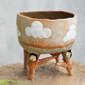 May include: A ceramic planter with a beige and brown gradient finish, featuring white embossed clouds, rain drops, and gold lightning bolts. The base consists of three brown tree-branch-shaped legs, each containing a small, round, yellow bird figure.