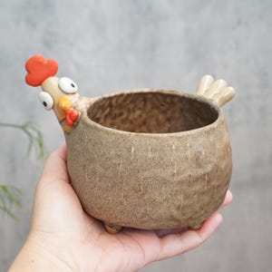 Puede incluir: Maceta de cerámica artesanal con forma de gallina. La maceta es de color marrón moteado con cresta y barbilla rojas, y grandes ojos saltones. Tiene una pequeña cola y tres patitas.