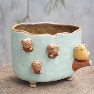 Puede incluir: Una maceta de cerámica azul claro con un borde ondulado y tres pequeñas decoraciones de nido tejido. Una pequeña figura de pájaro amarillo está posada en una rama en el lateral. La maceta tiene tres patas pequeñas.