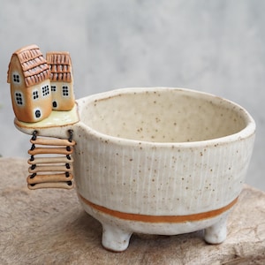 May include: A speckled off-white ceramic planter with three short legs and a horizontal orange band near the base. Attached to the rim is a miniature scene featuring two small tan houses with brown roofs and a hanging ladder made of clay rungs.