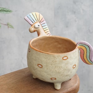 Puede incluir: Una maceta de cerámica caprichosa con forma de caballo. La maceta es de color beige con lunares marrones y una melena y cola de arcoíris. Tiene tres patas pequeñas y un cuerpo redondo, perfecto para plantas pequeñas.