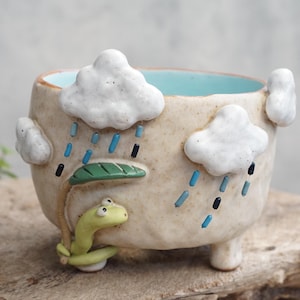 May include: A beige ceramic planter with three small feet, featuring raised white clouds, blue and black rain streaks, and a small green worm holding a green leaf as an umbrella. The interior of the round pot is glazed in a light blue color.