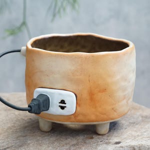 Puede incluir: Una taza de cerámica única con exterior beige y un interior marrón oscuro. La taza presenta un diseño de cable y enchufe negro, con un detalle de enchufe blanco. Se apoya sobre tres pequeños pies.