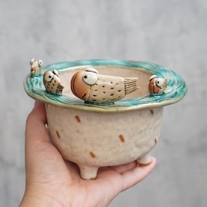 May include: A ceramic planter with a light beige body and a turquoise rim. The rim features a wave design and several fish figurines. The planter has three small feet and is decorated with brown spots.