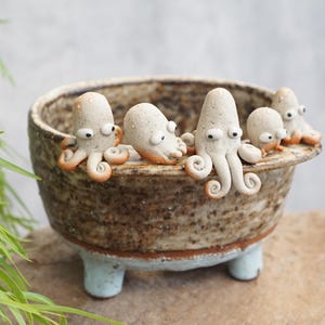 May include: A speckled brown ceramic bowl with a textured, rustic finish and three light blue feet. Five small, light-colored octopus figures with dark eyes and curled tentacles are perched along the rim of the bowl, facing outward.