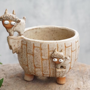 May include: A speckled beige ceramic planter with a brick-like pattern on the exterior. Two small, sculpted goat figures with white eyes and orange faces are attached to the rim and side of the pot, which stands on three short, rounded feet.
