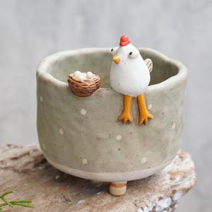 May include: A light green ceramic bowl with white polka dots, featuring a sculpted white chicken with a red comb and orange legs perched on the rim, next to a small brown nest containing three tiny white eggs, supported by three short feet.