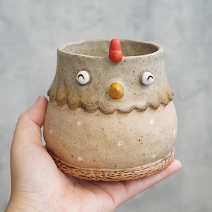 Puede incluir: Una taza de cerámica moteada con forma de pollo, cuerpo color canela claro, detalle de cuello festoneado, pico naranja, dos ojos redondos y una cresta roja. La base presenta un patrón texturizado y pequeños puntos blancos como acentos.