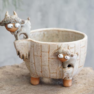 May include: A whimsical ceramic planter with a light brown, textured finish. The planter features three small legs and is adorned with two cartoonish goat figures with large eyes. The goats are light gray with brown accents.