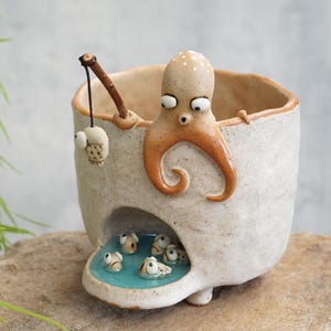 May include: A speckled off-white ceramic planter featuring a sculpted orange octopus holding a brown twig fishing rod with a small fish attached. A small arched opening at the base reveals a turquoise pool containing several tiny ceramic fish.